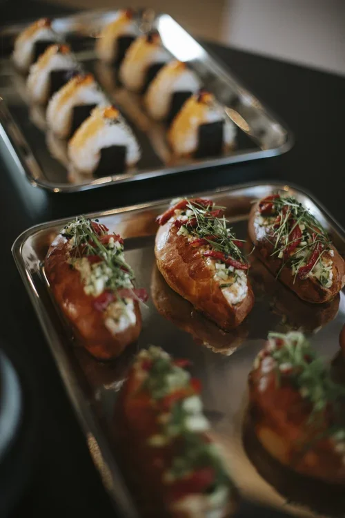 Tray of stuffed potato skins topped with microgreens and ketchup, with a blurred tray of sushi in the background.