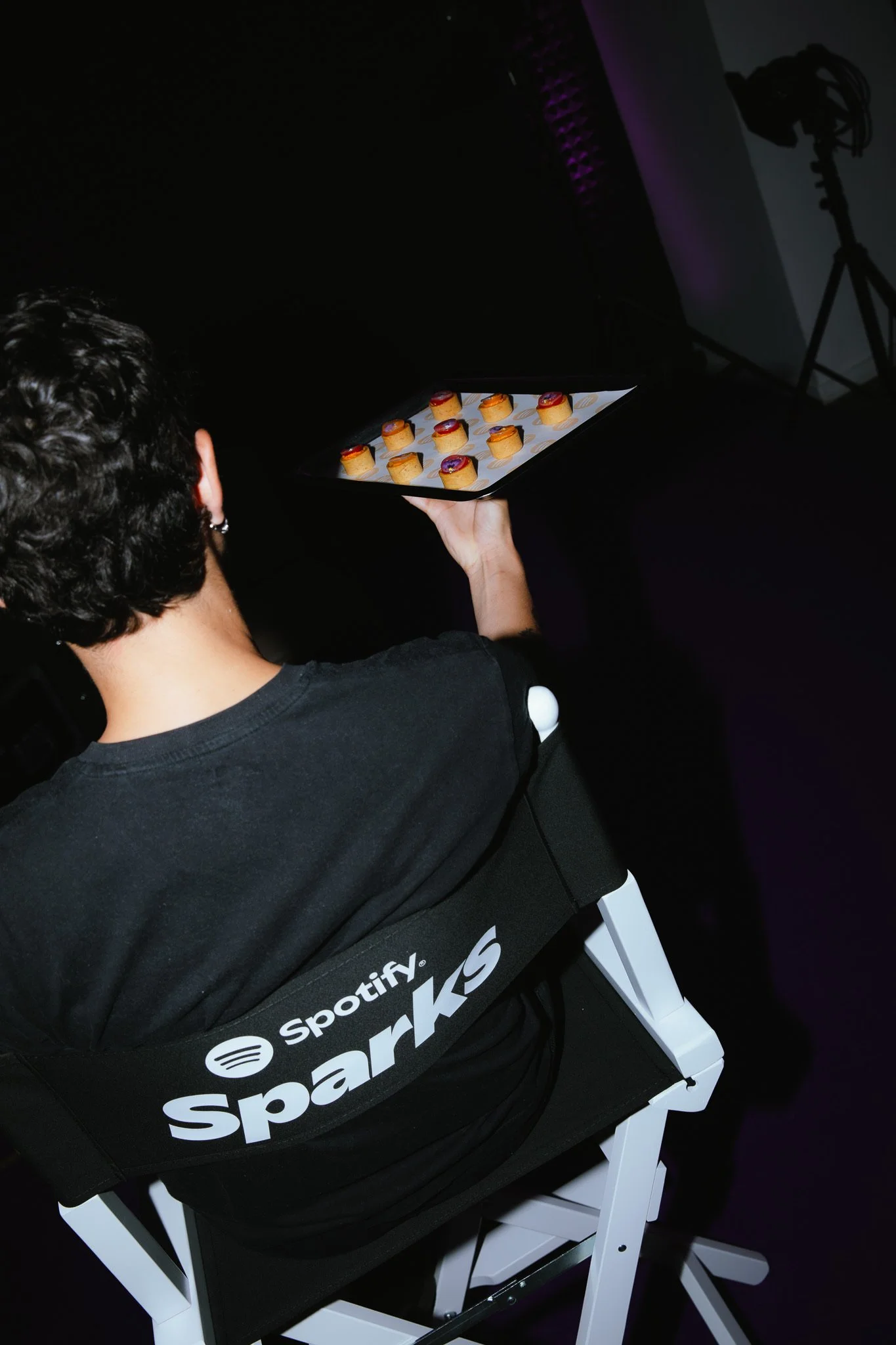 Person seated on a chair with Spotify SparkS branding, holding a tray of small desserts in a dimly lit room.