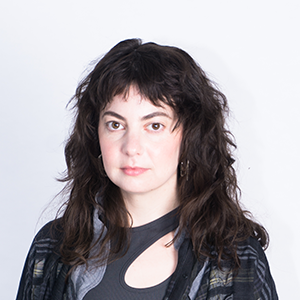 A young woman with wavy dark hair, wearing a dark striped jacket over a dark top, standing against a plain white background.