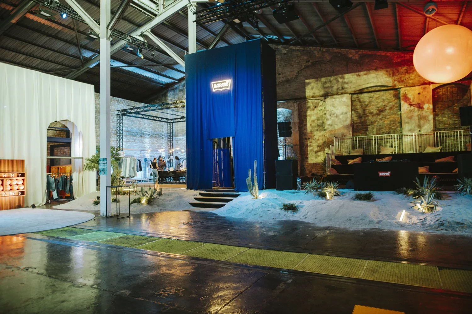 Indoor event space with a desert-themed stage, Decor includes sand, cacti, and desert plants, blue curtain backdrop with illuminated sign, modern lighting, and seating area.