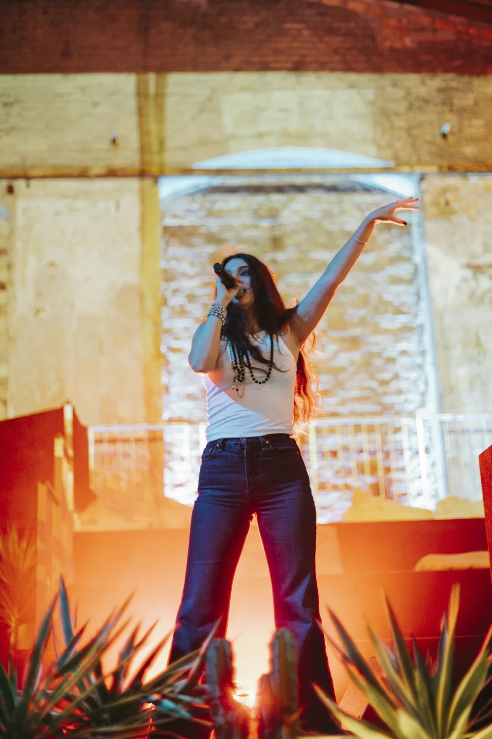 A woman singing into a microphone on stage with an exposed brick wall background and stage lighting.