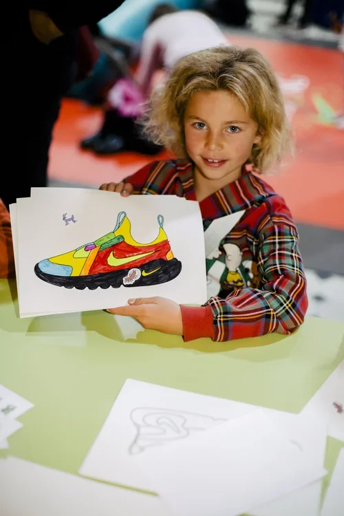 A young girl with curly blonde hair holding a drawing of a colorful sneaker, sitting at a green table in a room with other children.
