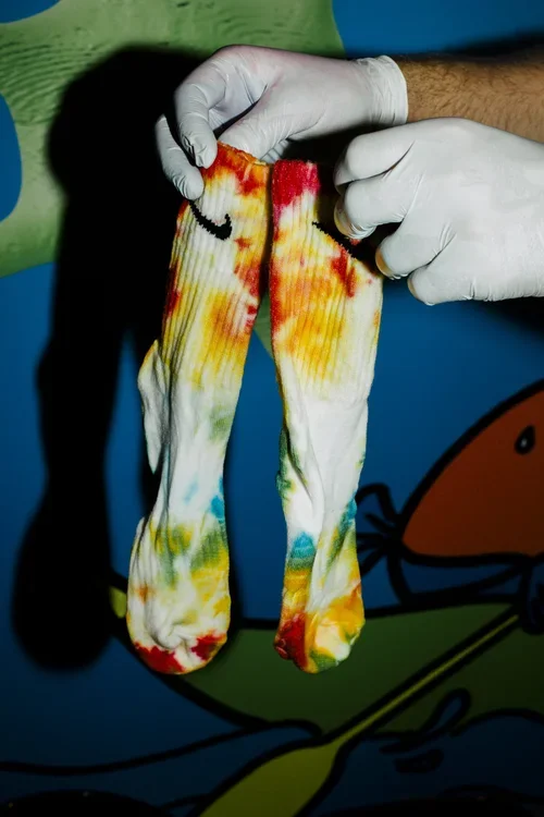 Person wearing gloves holding a colorful tie-dye pair of socks against a blue and green background.