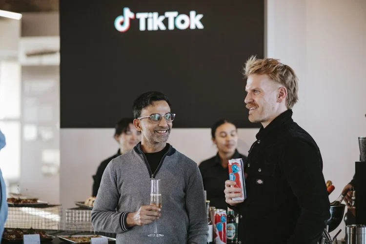 Two men smiling and talking at a social gathering with a TikTok logo on a large screen in the background.