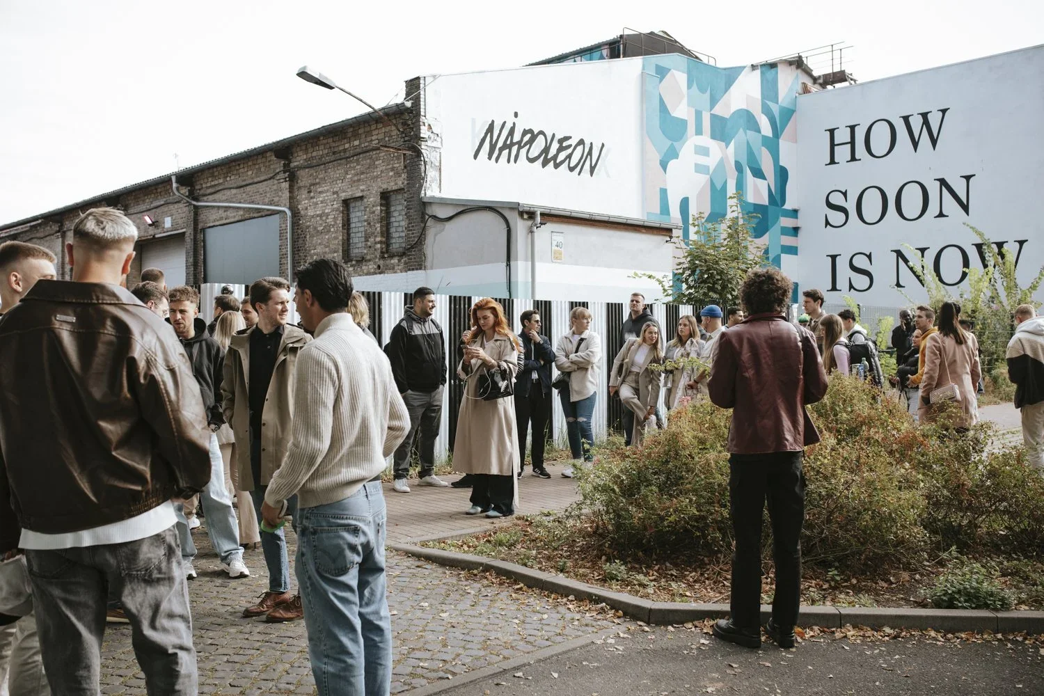 A group of people standing in line outdoors near a building with urban graffiti art and large signs that read 'NAIOLEON' and 'HOW SOON IS NOW.'