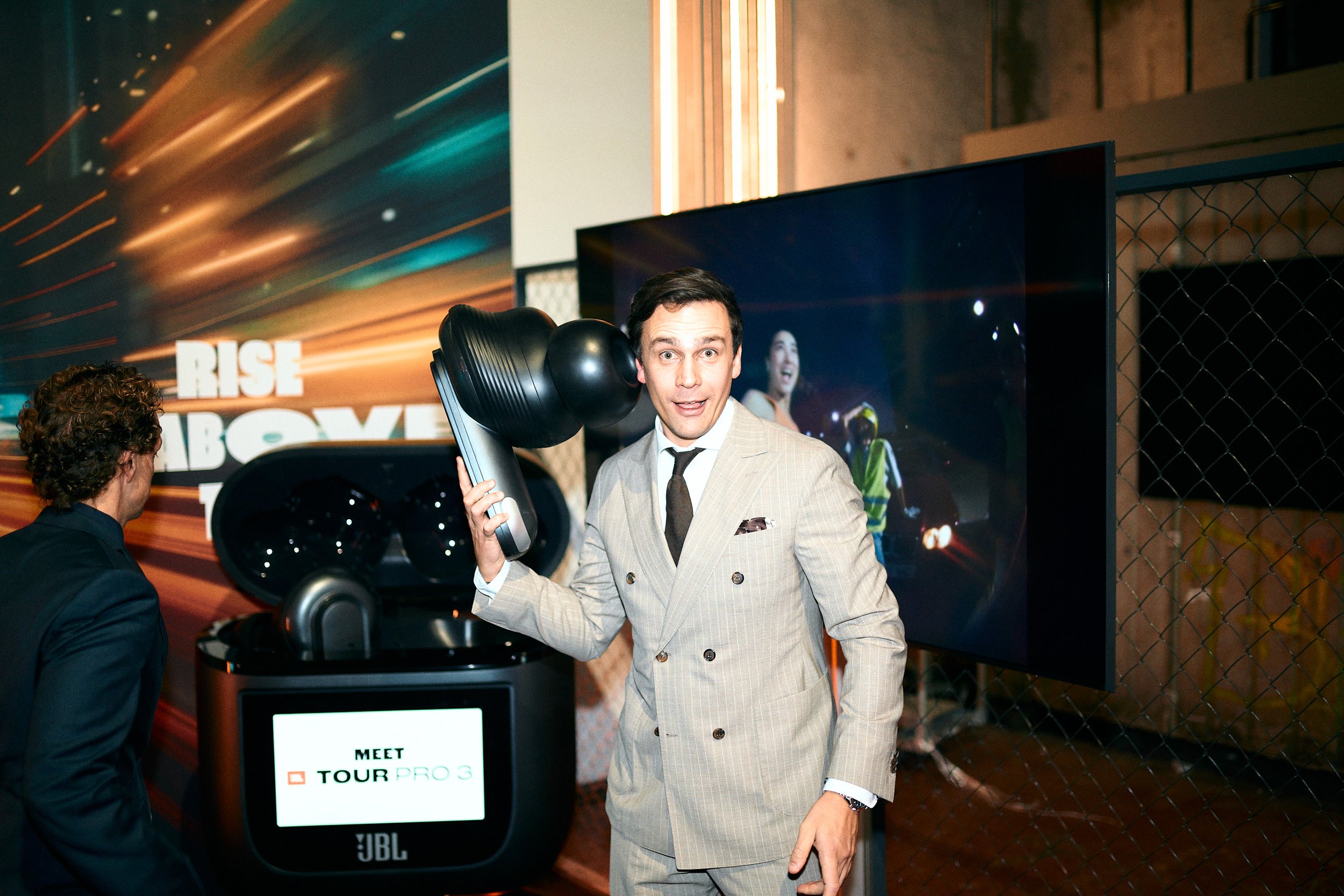 A man in a light-colored suit holding a virtual reality headset with headphones at an event, with a large screen and promotional background behind him.