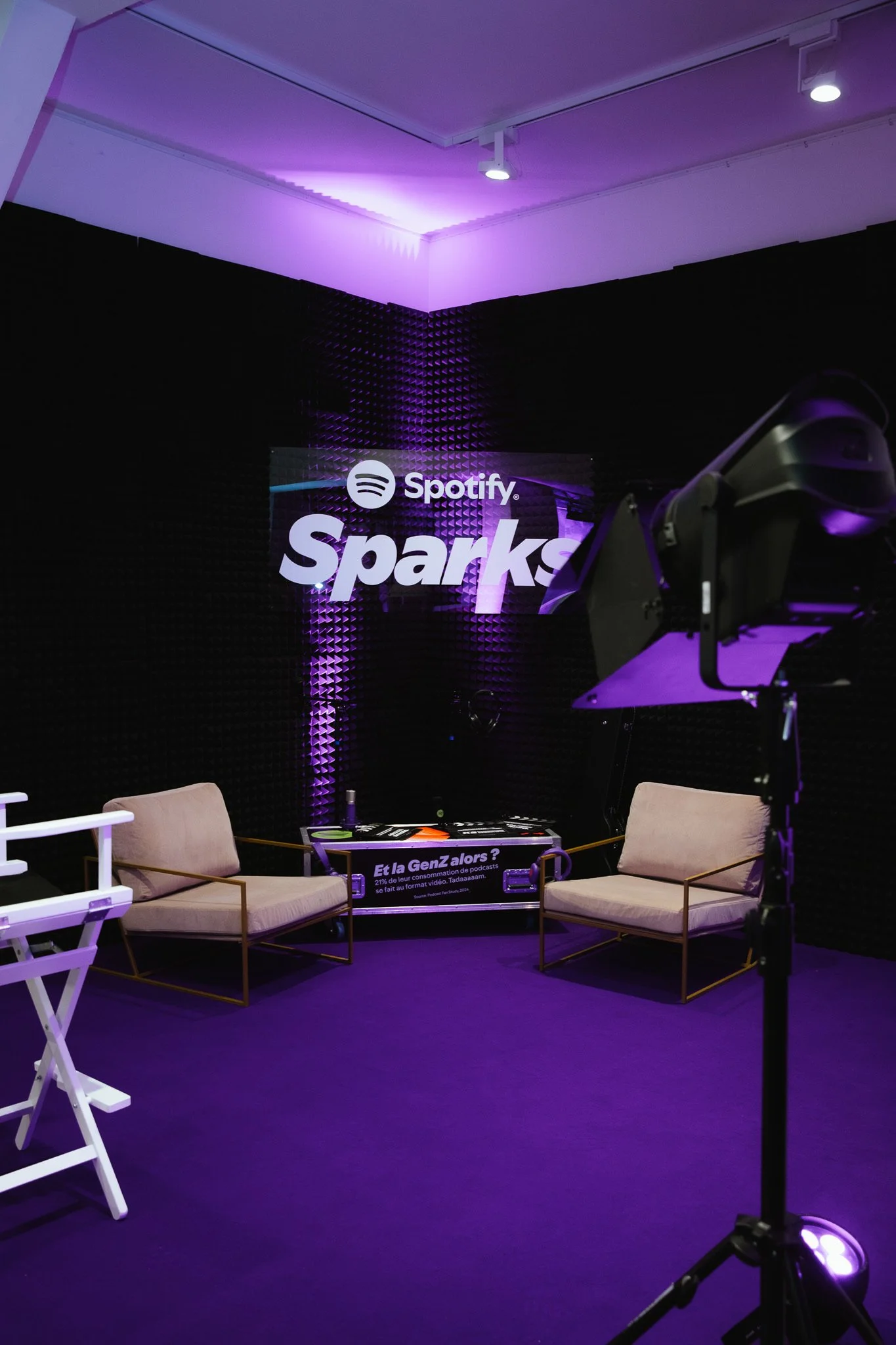 A recording studio with black acoustic foam walls, purple ambient lighting, a sign that says Spotify Sparks, two beige armchairs, a DJ turntable, and professional studio lighting equipment.