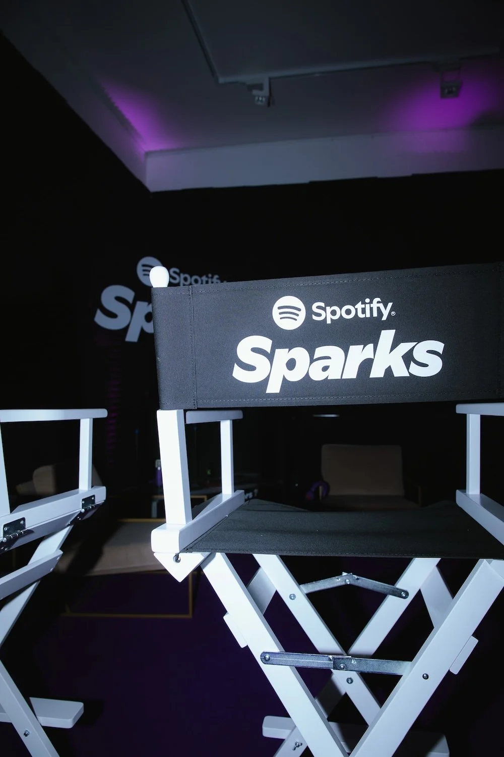 A director's chair with a black fabric seat and backrest labeled 'Spotify Sparks' in white letters, set against a dark background with additional chairs and purple lighting.