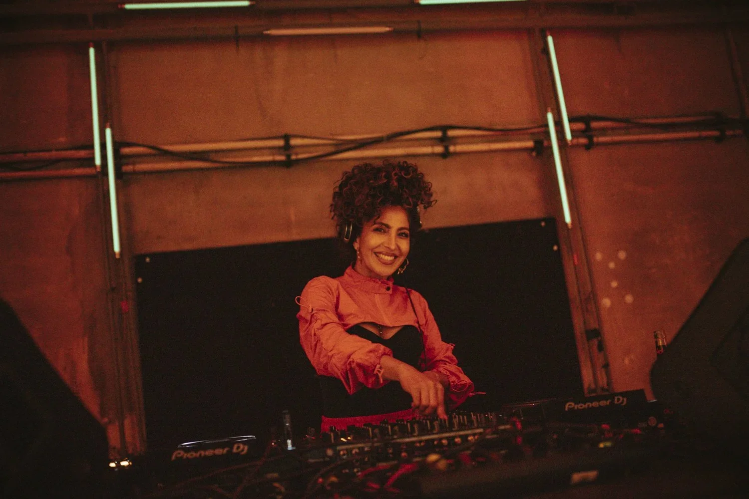 A woman with curly hair, wearing a red and black outfit, smiling while DJing at a nightclub. She is standing behind a Pioneer DJ setup with colorful lighting in the background.
