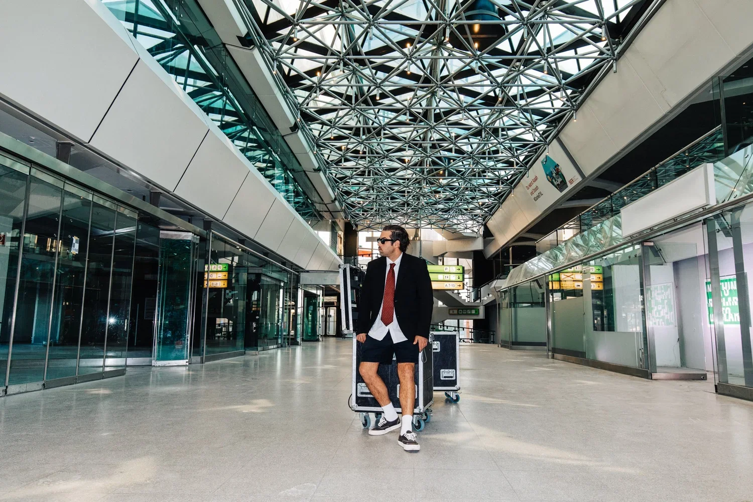 A man in a suit jacket, shorts, and sneakers stands in an airport with a trolley, in front of glass doors and a high, geometric glass ceiling.