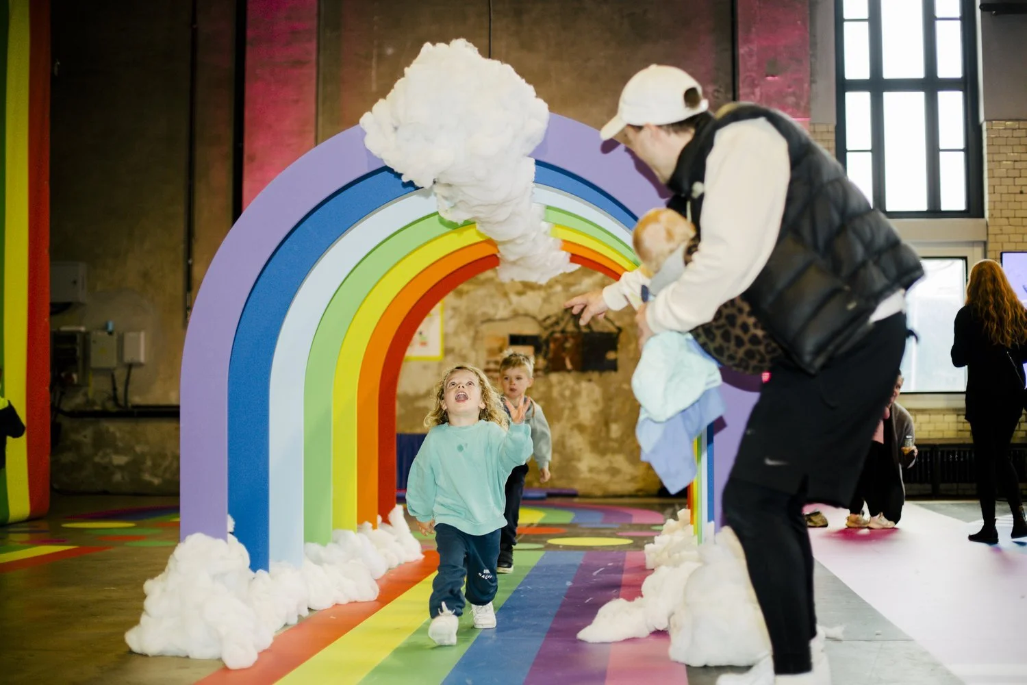 Children running through a rainbow-themed archway decorated with fluffy white clouds in an indoor play area.