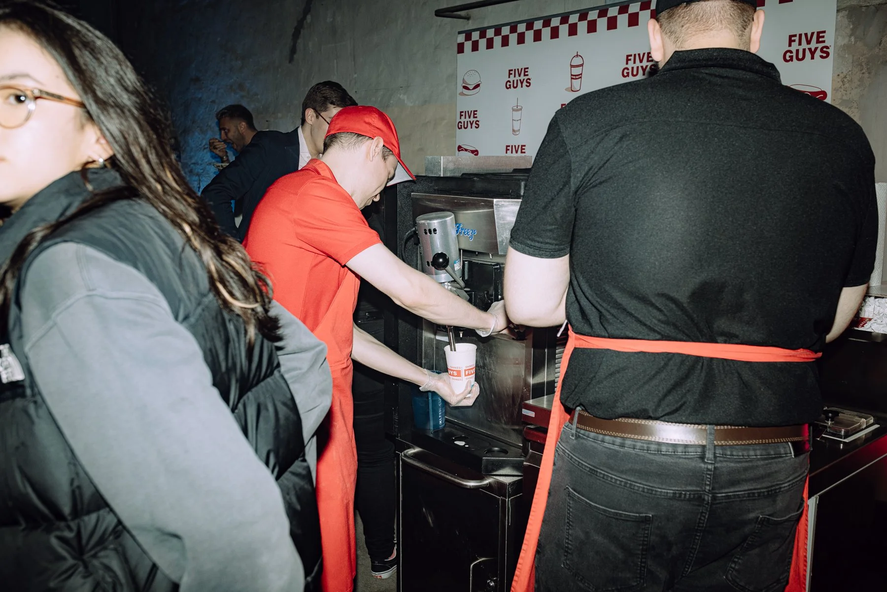 People lining up at a Five Guys fast-food restaurant, with the staff preparing a drink behind the counter.