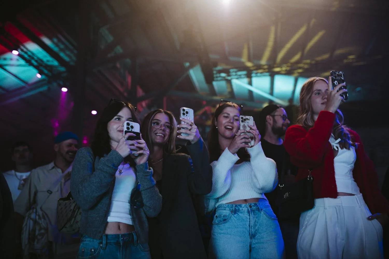 Group of young people enjoying a concert or event, taking photos with smartphones, in a dimly lit space with colorful lights.