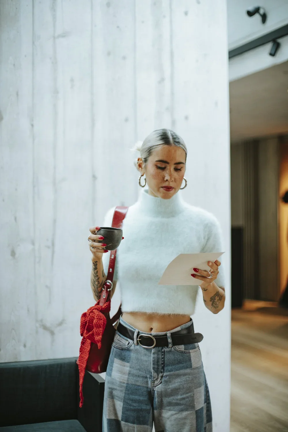 A young woman with platinum blonde hair, tattoos on her arms, and hoop earrings is reading a piece of paper. She is holding a black mug in her left hand, wearing a white cropped turtleneck sweater, high-waisted distressed jeans, and a black belt. She