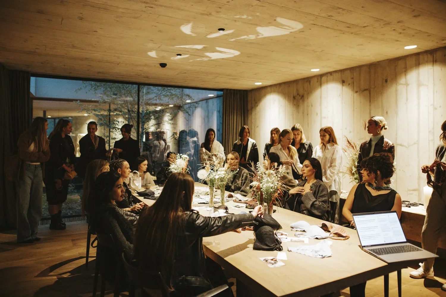 Group of women gathered around a large wooden table with flowers and papers, some women are standing and some are sitting, in a modern well-lit room with big windows.