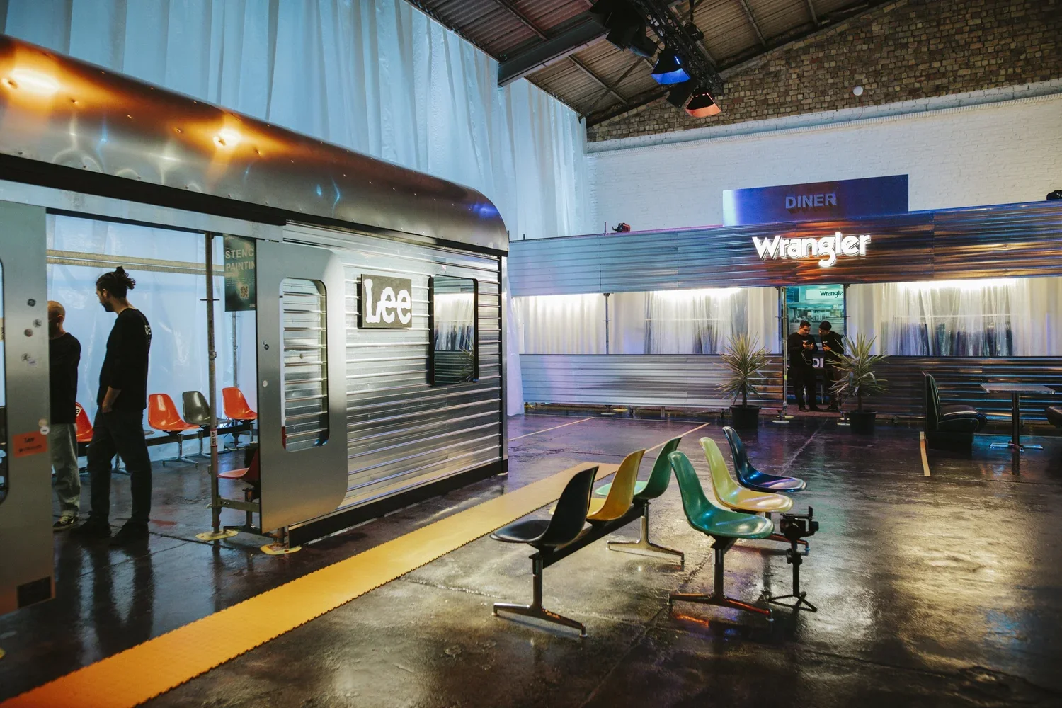 Interior of a modern, industrial-style diner with colorful chairs, a vintage trailer labeled "Lace," and illuminated signs for "Wrangler" and "Diner."
