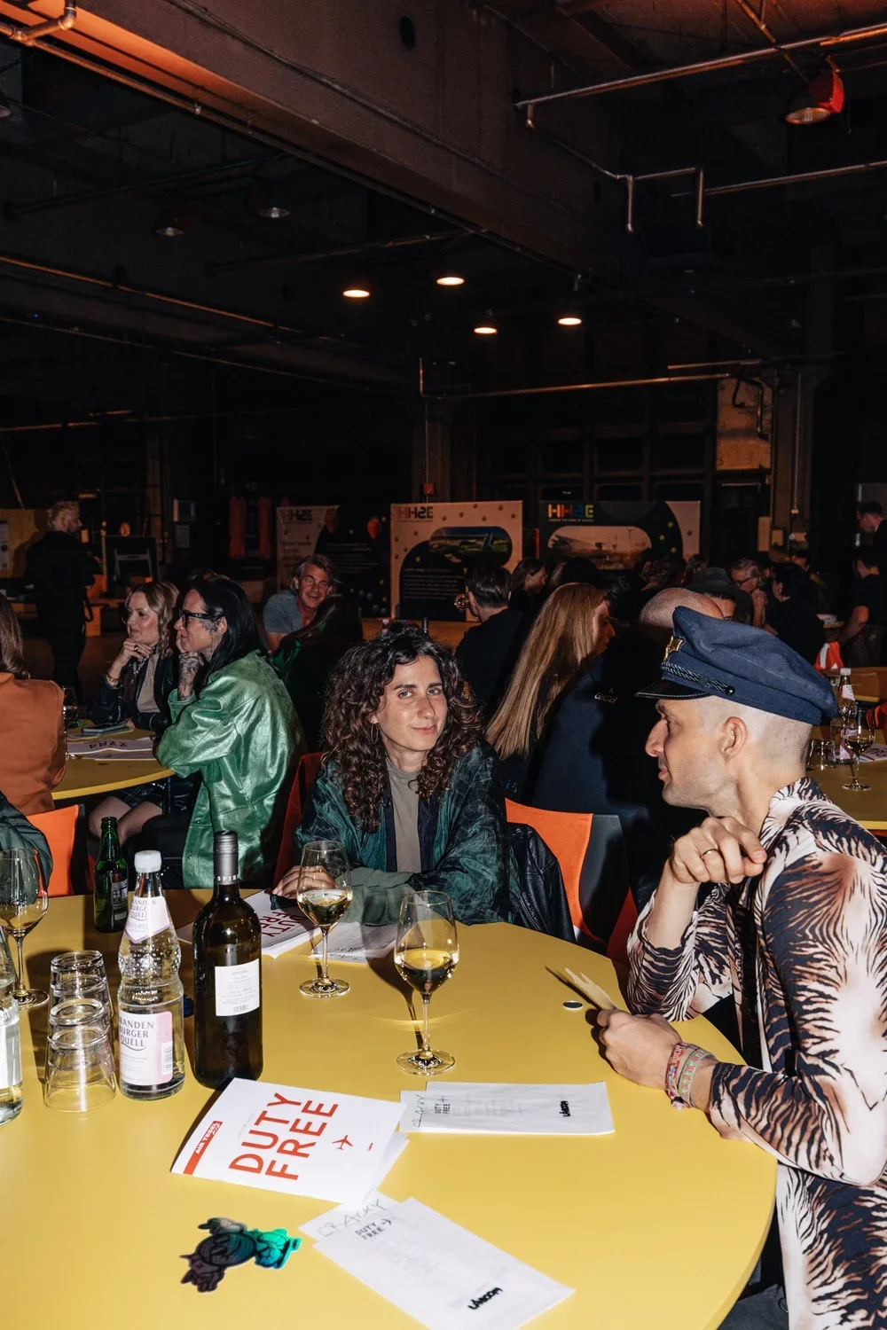 People sitting at a yellow table in a dimly lit venue, some with wine glasses, and a man in a tiger-print shirt with a blue cap, showing a menu or card.