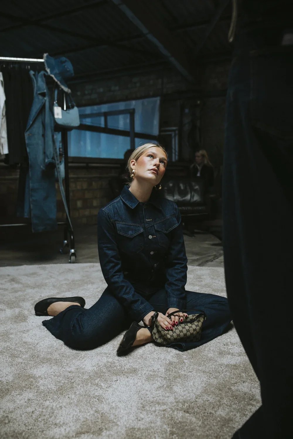 A young woman sitting on the floor of a clothing store, holding a designer handbag, wearing a denim jacket and jeans, with blonde hair and gold earrings, looking upwards amidst racks of clothes and a brick wall in the background.