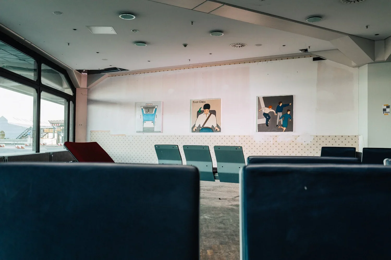 Empty airport seating area with chairs facing a white wall, decorated with three framed posters depicting airplane travel and passengers. Large windows on the left show parts of the airport tarmac outside.