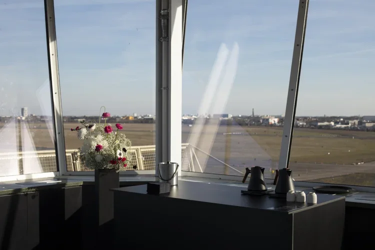 View from an airport terminal window showing an airfield with runways, a flower vase with pink and white flowers, and some coffee pots on the counter.