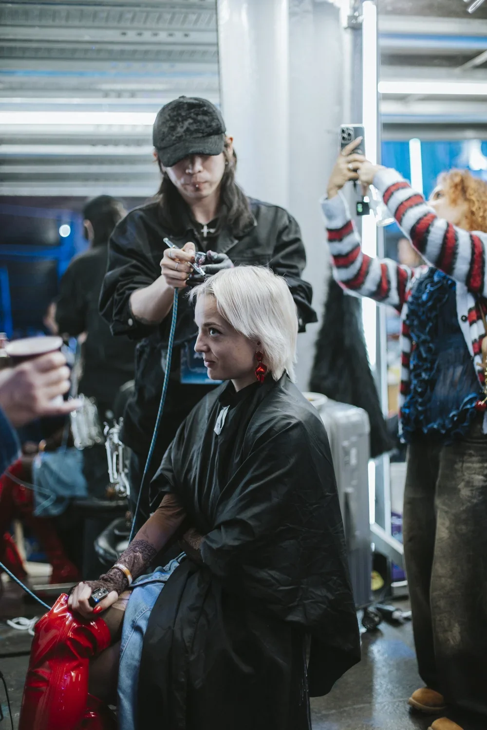 A woman with platinum blonde hair getting a haircut by a hairstylist in a busy salon or backstage area, with other people and equipment in the background.