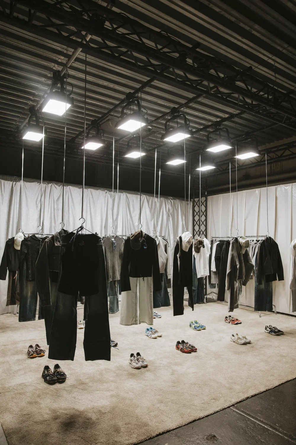 Clothing display with hanging jackets, pants, and shoes on a beige carpeted floor, in a large indoor space with a black ceiling and bright overhead lights.