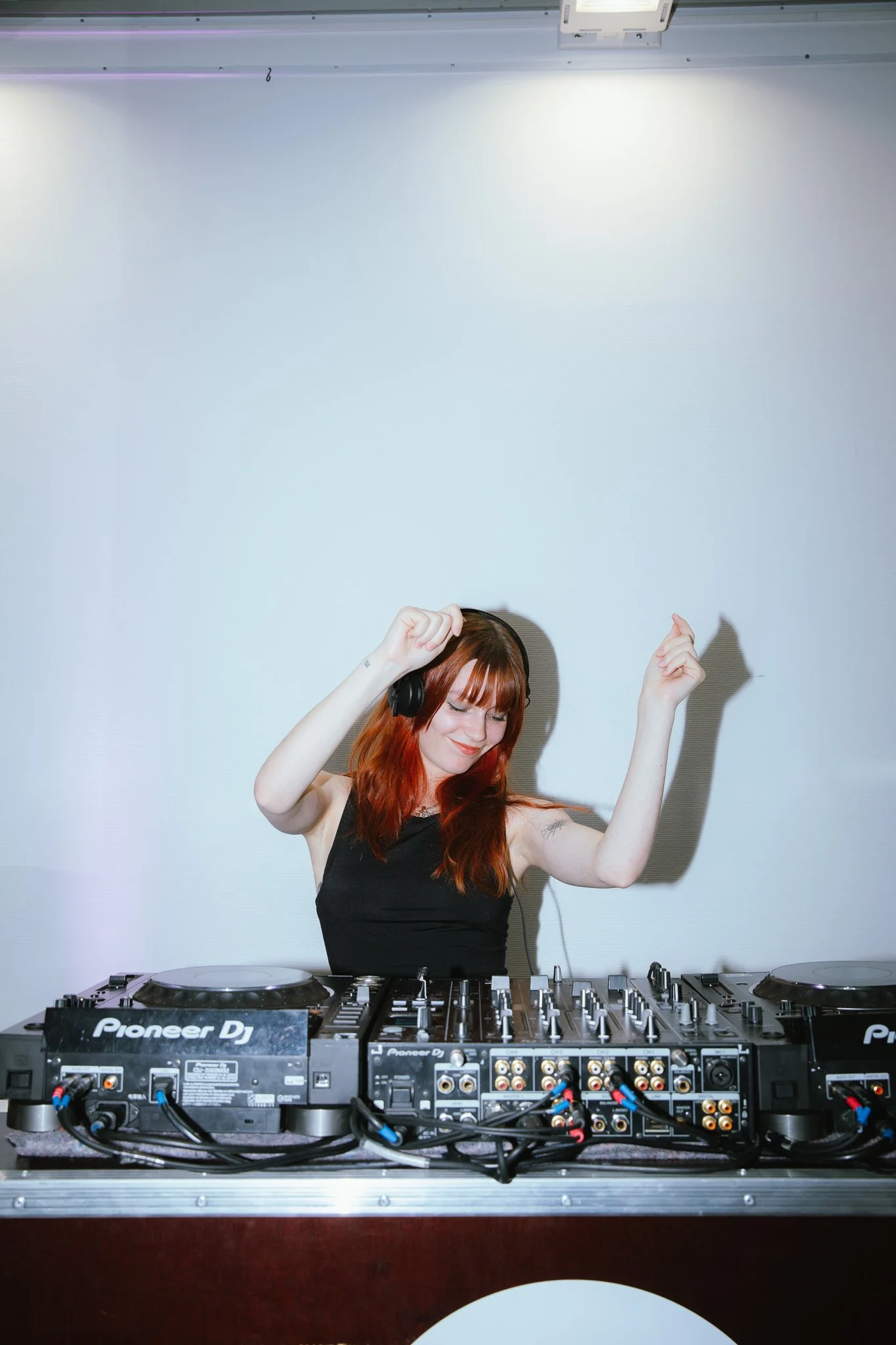 A young woman with red hair and black headphones dancing behind DJ equipment in a studio or club setting.