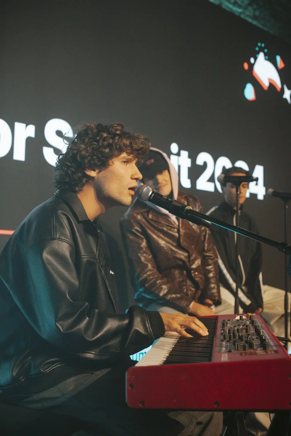 Young man with curly hair wearing a black leather jacket playing a red keyboard and singing into a microphone, with two other young men in the background, one wearing a hoodie and the other in glasses and a dark jacket, in front of a dark background 