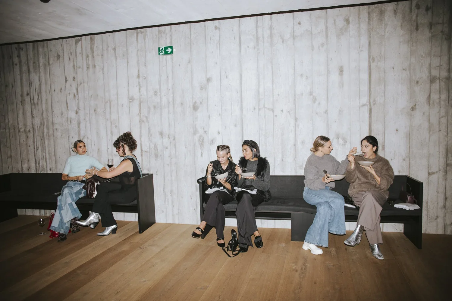 Five women sitting on benches against a gray wall, with some holding wine glasses and eating from bowls, in an indoor setting.