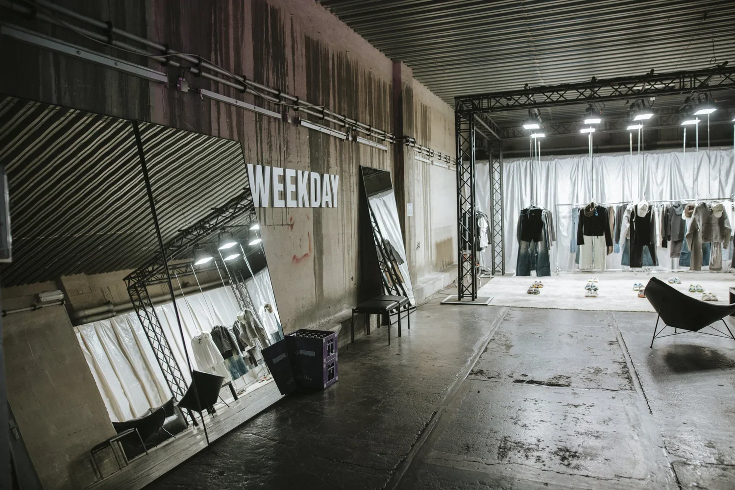 Indoor fashion display with clothing racks, mannequins, and stage area, sign reading 'WEEKDAY', mirrors, and chairs in an industrial-style space.