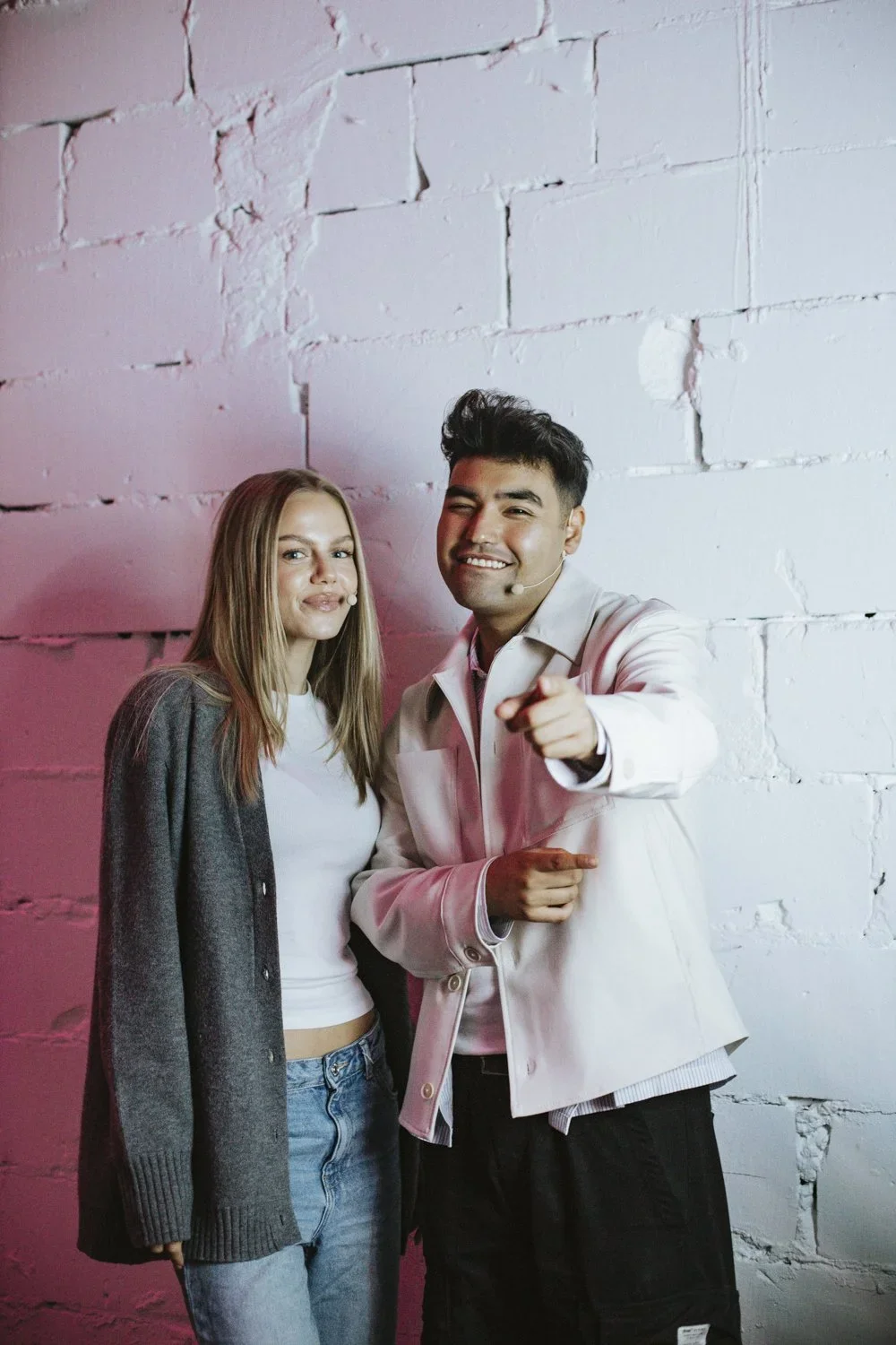 A young man and woman standing together against a white brick wall, both wearing headsets with microphones, smiling, and the man pointing at the camera.
