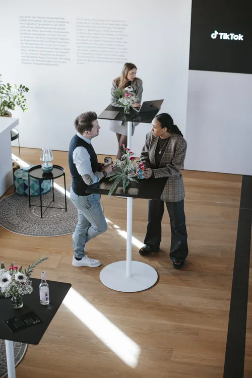 Two people talking at a high-top table with flowers and drinks, a woman standing nearby with a laptop, and a woman on the side in a blazer, in a modern room with TikTok logo on the wall.