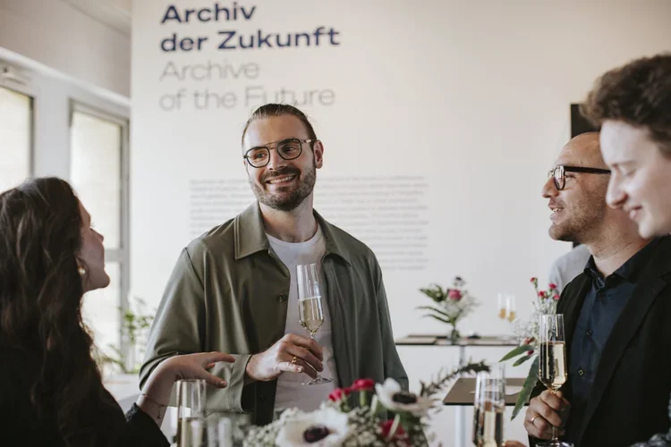 People at a social event holding glasses of champagne, smiling and talking in a room with minimalistic decor and a sign that reads 'Archive of the Future' in German and English.