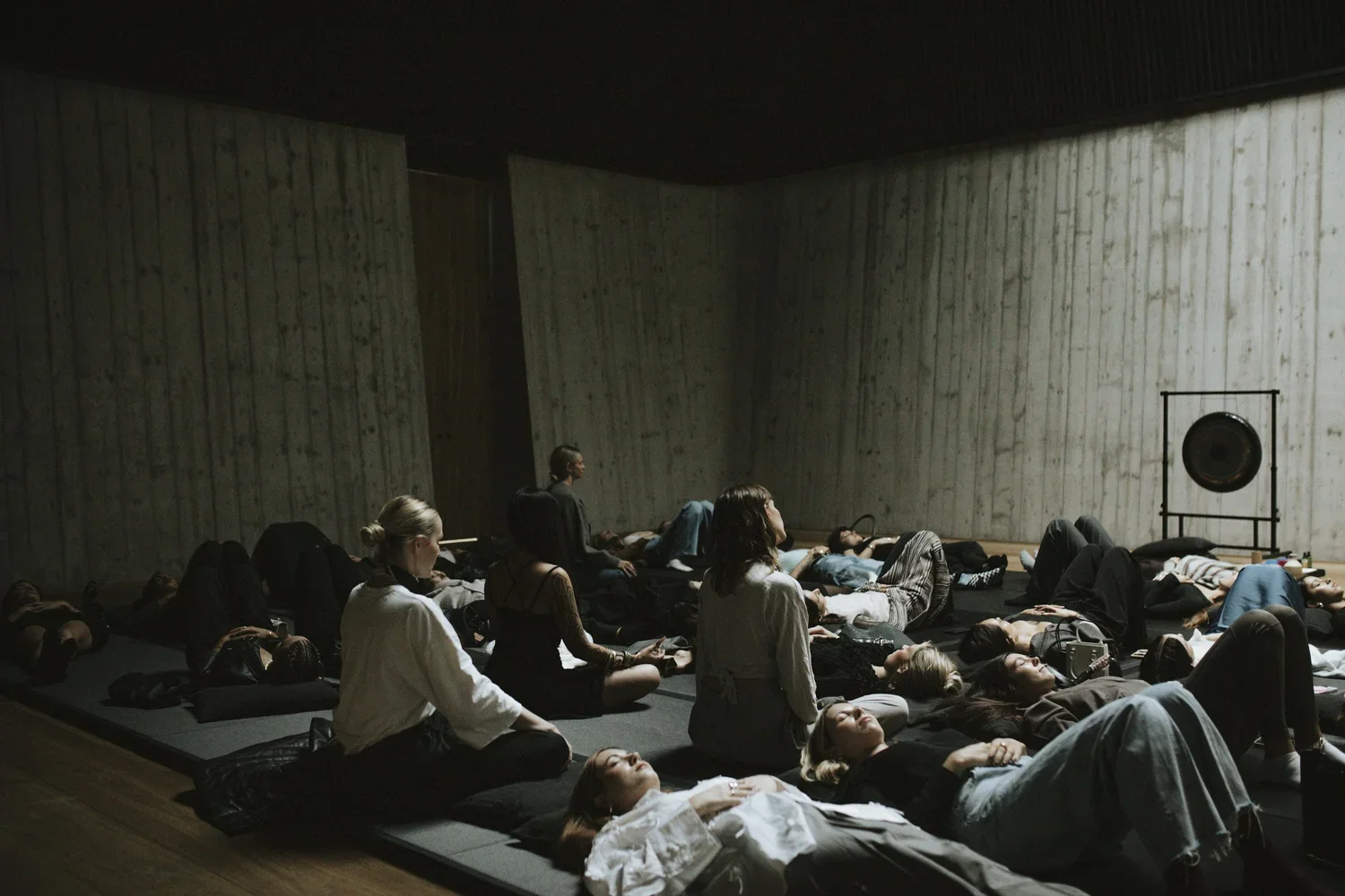 People lying down on mats in a dark meditation or yoga space with a gong in the background.