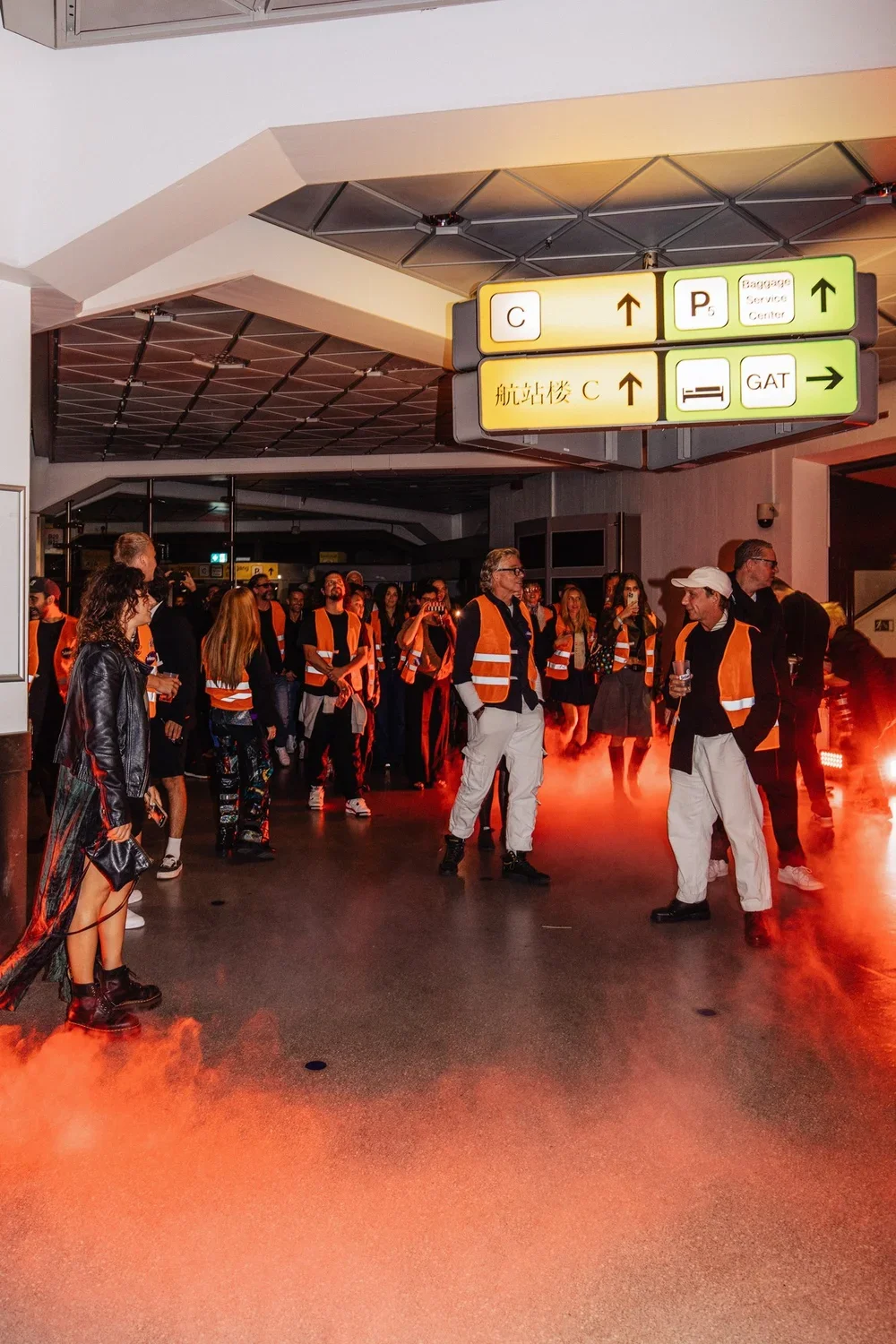 A group of people, some wearing orange vests, gathered in an airport terminal with red fog or smoke on the floor. Overhead signs indicate directions to gates, parking, baggage service, and a GAT area.