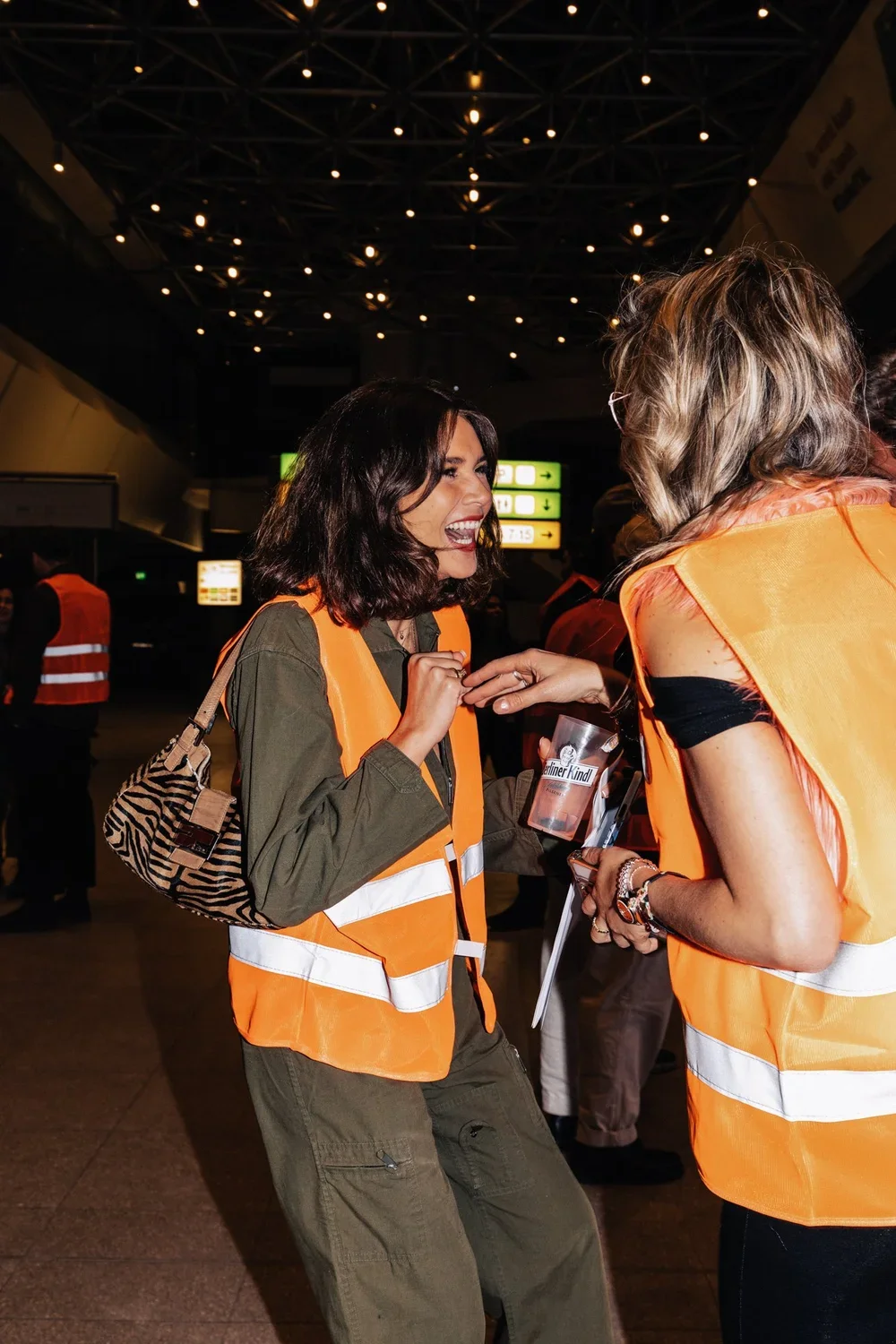 Two women in orange safety vests smiling and talking at an airport terminal, with airport signage and other people in the background.