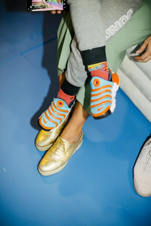 Child wearing colorful athletic shoes with orange and blue stripes, gold slip-on shoes, gray and black pants, and a gray sweatshirt, sitting on a blue floor and holding a smartphone.