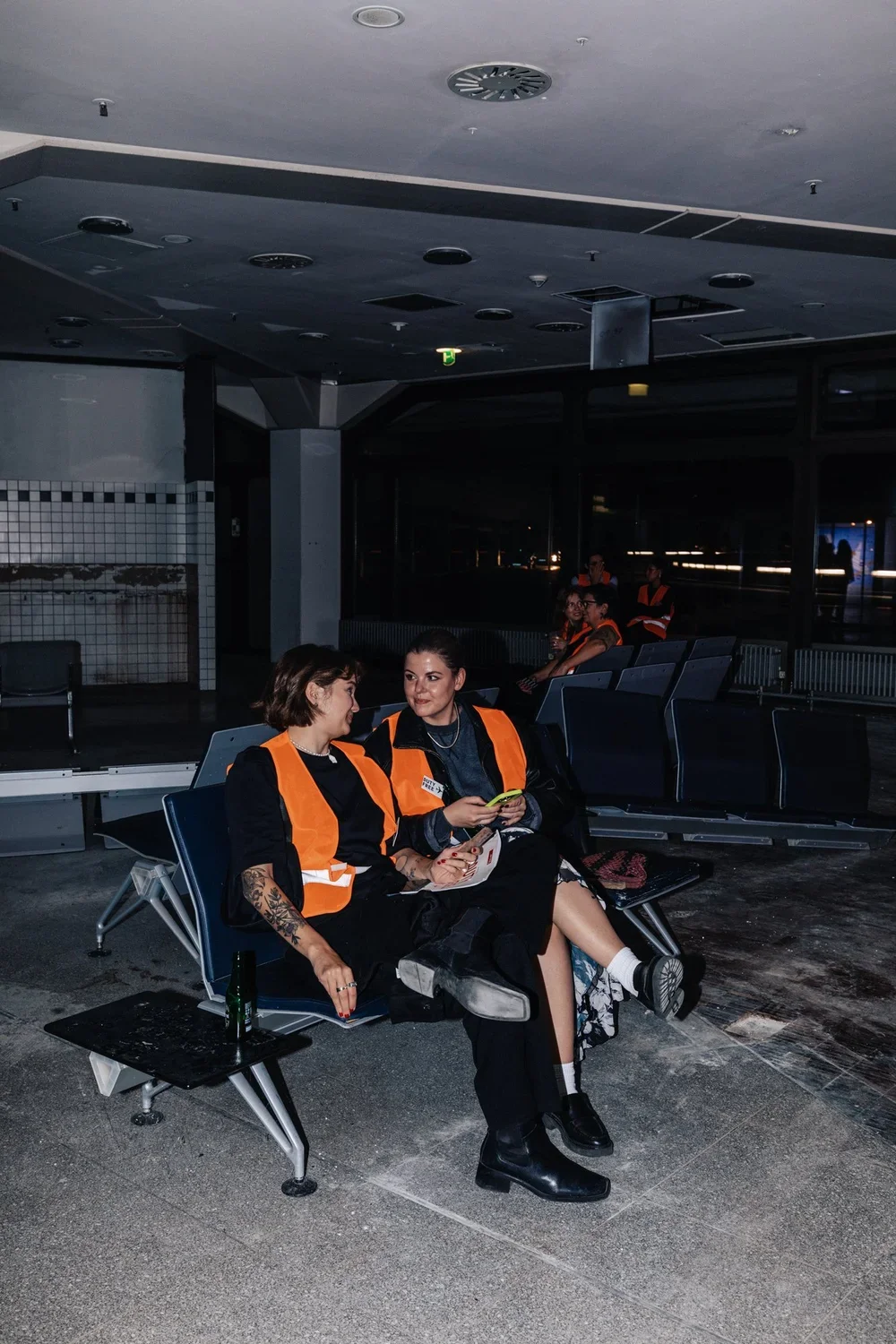 Two women wearing orange safety vests sitting and talking on airport benches, with other women in safety vests in the background indoors at night.