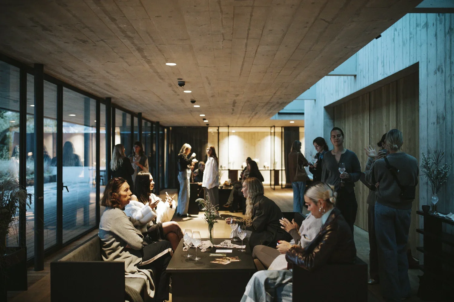 Group of people socializing at an indoor gathering with modern decor, large windows, and ambient lighting.