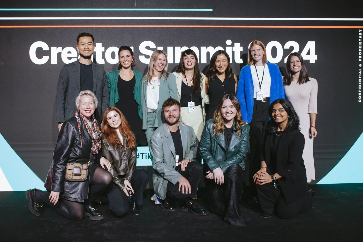 A diverse group of 13 people, mostly smiling, posing for a photo on a stage in front of a large screen that reads 'Creators Summit 2024' during an event.