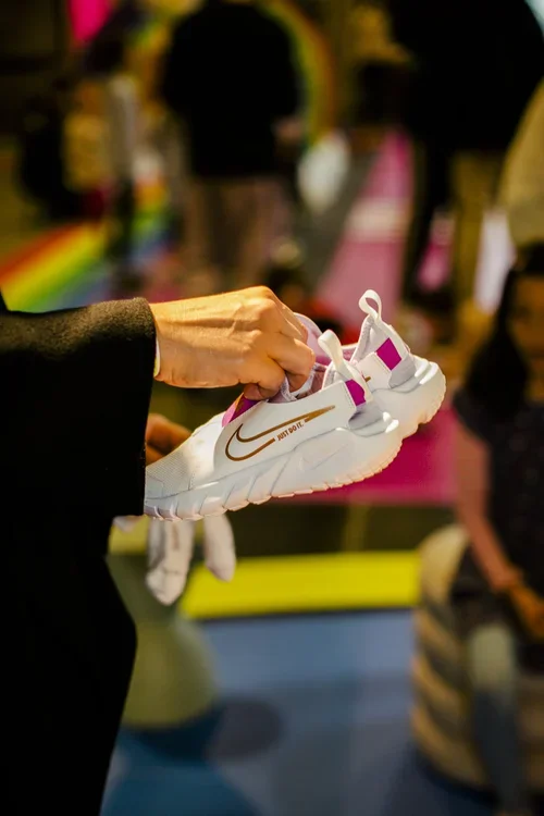 Person holding a white Nike athletic shoe indoors with colorful blurred background and other people.