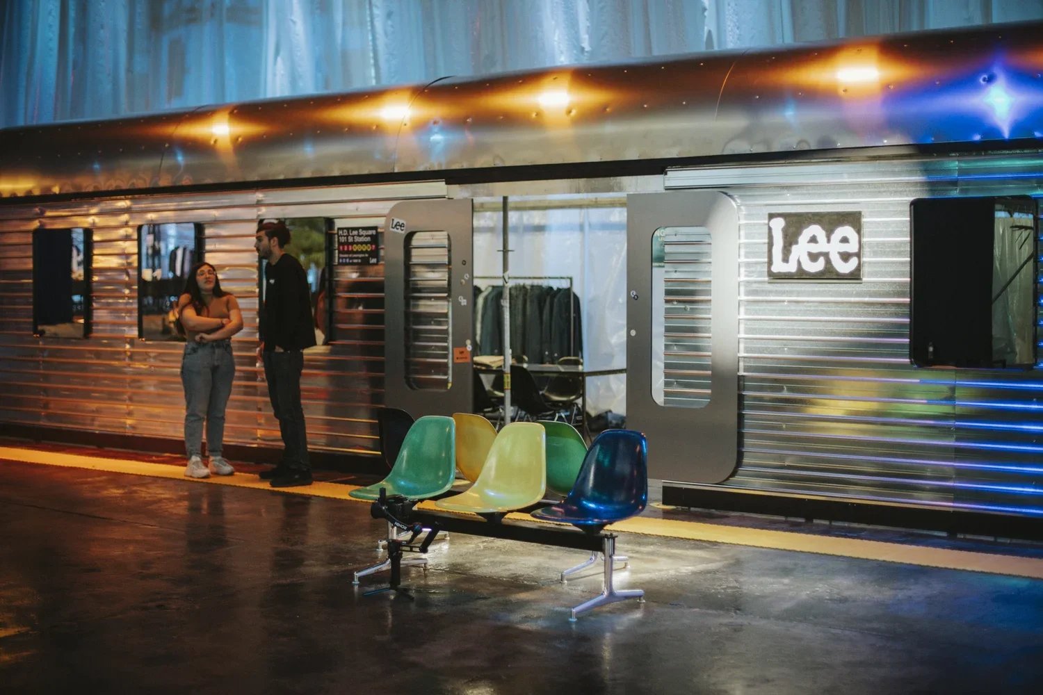 People standing and talking on a train platform at night with colorful chairs in the foreground and a silver train with the sign 'Lee' in the background.