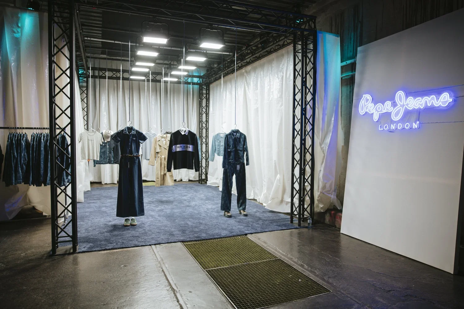 Clothing display inside a Pepe Jeans London store with a black metal frame, hanging garments, and a neon sign on the right side that reads 'Pepe Jeans London'.