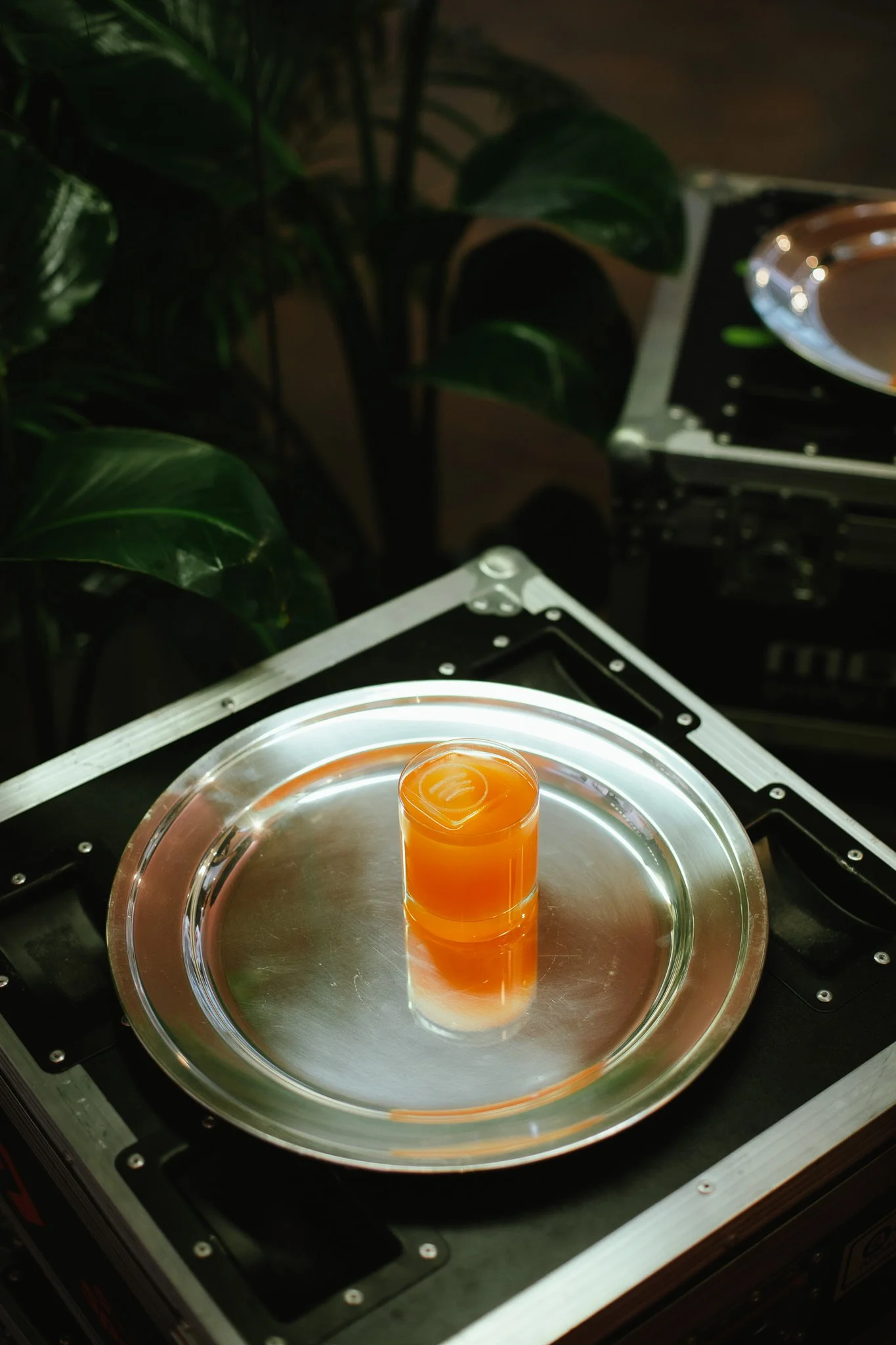 A drink with orange color in a transparent glass on a silver tray, placed on a black turntable or DJ equipment, with green leaves in the background.