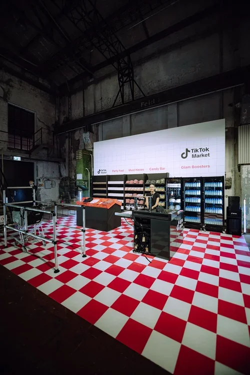 Empty indoor marketplace with a TikTok Market booth featuring shelves of products and a large LED screen display, red and white checkered flooring, and industrial ceiling.