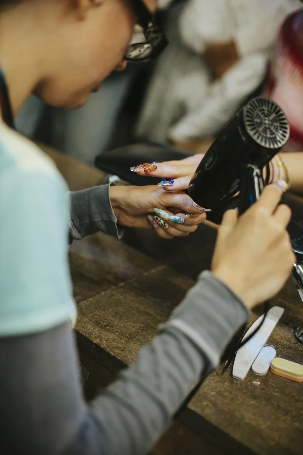 Person drying nails with a hairdryer in a salon, with various nail tools on the wooden table.