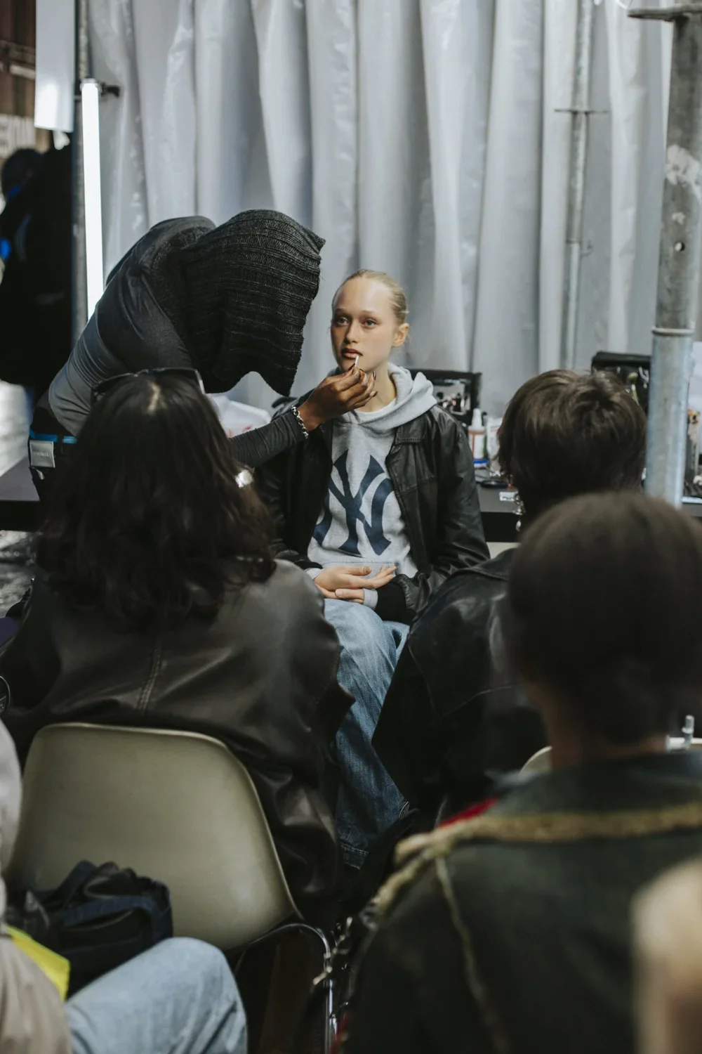 A woman with a large black hood is applying makeup to a young woman sitting with her hands in her lap during a backstage or salon setting, surrounded by other seated individuals, with gray curtains in the background.