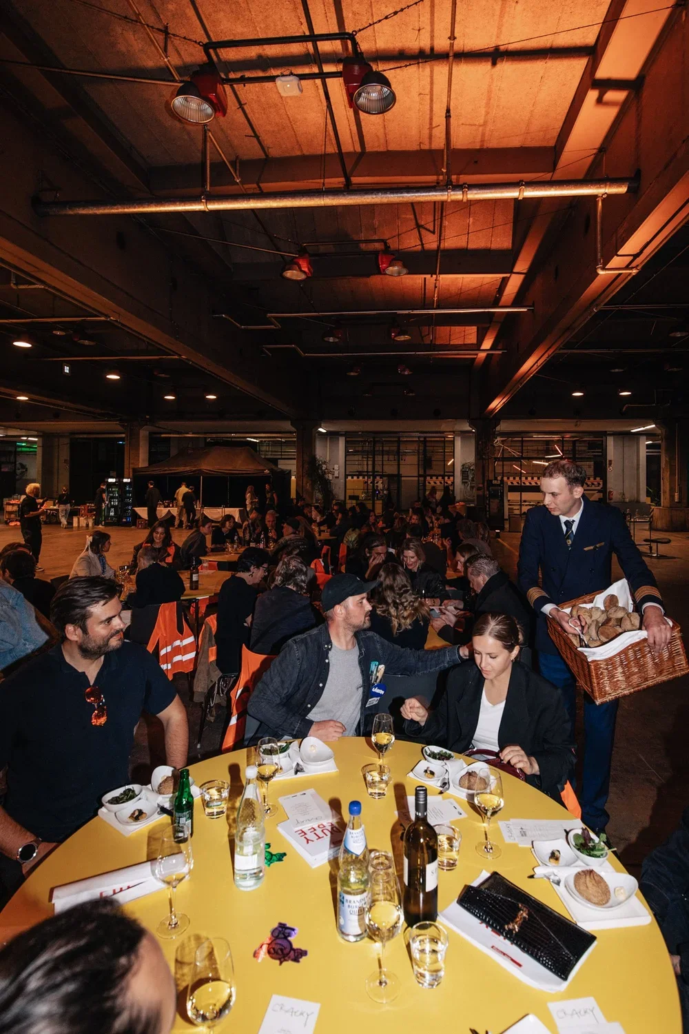 People seated at a round table enjoying a dinner, with wine glasses and plates of food, in a large industrial-style venue with high ceilings and exposed pipes.