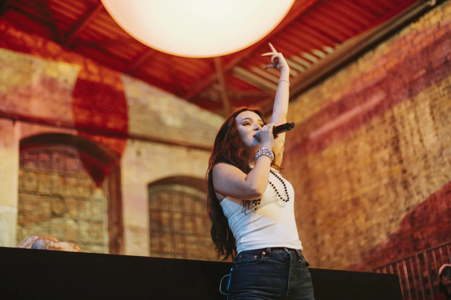 A woman with long, wavy dark hair singing into a microphone on stage, raising one hand in the air. She wears a white tank top, black jeans, and jewelry. The background features a brick wall and warm lighting in a rustic venue.