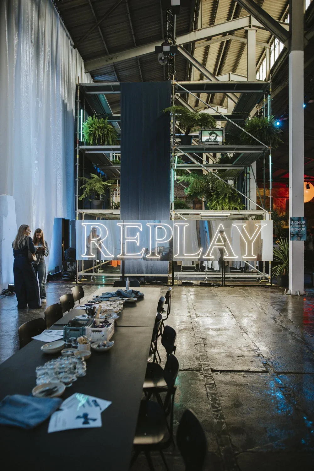 Indoor event space with a large illuminated sign that reads 'REPLAY' in front of a multi-storey scaffolding structure decorated with plants. There are tables with plates and glasses, and two women are standing and talking near a large white curtain w